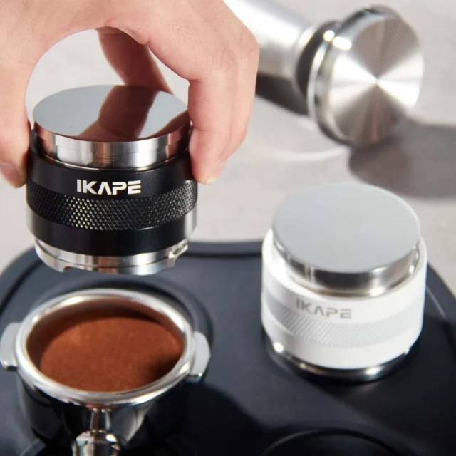 Hand hält das schwarze Werkzeug an der flachen Seite über einen Siebträger mit Kaffeemehl, dahinter die weiße Version