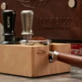 Schwarzer und silberner IKAPE Tamper in der Buchenstation, ein Siebträger mit gehäuftem Kaffeemehl im Schlitz