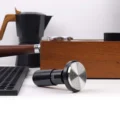 Tamper liegt auf der Seite auf weißer Arbeitsfläche, Basis mit Ringen nach außen, dahinter Espressokocher und Holzbox