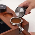 Hand senkt den Tamper auf einen Siebträger mit geebnetem Kaffee in einer Holzbox