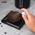 Hand sprüht mit der weißen Flasche über die Kaffeebohnen in der Schale auf der Waage, dahinter eine Bohnendose