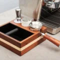 Bottomless-Siebträger mit Dosiertrichter auf einem zweifarbigen Abklopfbehälter aus Holz, mit Tamper und Distributor