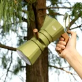 Hand hält einen grünen Espressokocher am Griff in Holzoptik im Freien, unter den Ästen eines Baums