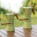 Zwei grüne Espressokocher, ein kleinerer und ein höherer, auf einem Holzlattentisch im Freien
