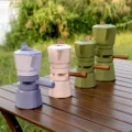 Violetter, rosa und zwei grüne Espressokocher auf einem Holztisch im Freien, dahinter Gras und Sträucher