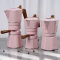 Drei rosa Espressokocher in einer Reihe auf grauem Marmor, zwei davon mit geschlitztem Mittelring und Seitengriff