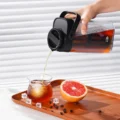 Hand gießt Cold Brew durch die dichte Ausgusskappe des schwarzen Krugs in ein Glas mit Eis, Grapefruit auf dem Tablett
