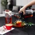 Hand gießt Cold Brew aus dem schwarzen Krug in ein Glas mit Eis, dahinter der braun-cremefarbene Krug