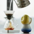 Gelbe Tasse umgedreht auf einer blauen Verlaufstasse, dahinter unscharf ein Handfilter-Aufguss