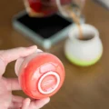 Hand hält eine rote Tasse umgedreht und zeigt das IKAPE Logo im Bodenring, dahinter eine hellgrüne Tasse