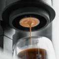 Espresso mit Crema läuft aus dem schmalen Siebboden unter einem schwarzen Brühgerät mit gerändeltem Ring