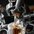 Nahaufnahme: Espresso fällt aus einem Bottomless-Siebträger in ein Glas mit Eis, daneben etwas Dampf