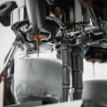 Espresso läuft aus einem Bottomless-Siebträger in ein Glas Milch, rechts unscharf eine zweite Brühgruppe