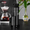Schwarze IKAPE Mühle auf einem dunklen Holztisch neben einem gläsernen Handfilter und einer Pflanze in einer Vase