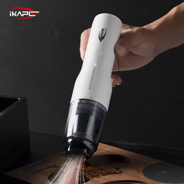 IKAPE Kaffee Staubsauger in Weiß, in einer Hand, saugt verschüttetes Kaffeemehl von einem Holztablett
