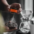 Orange Sprühflasche besprüht Kaffeebohnen in einer kleinen Schale in einer Hand, dahinter ein Pour-over-Dripper