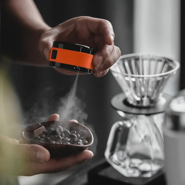 Orange Sprühflasche besprüht Kaffeebohnen in einer kleinen Schale in einer Hand, dahinter ein Pour-over-Dripper