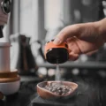 Orange Sprühflasche besprüht eine Holzschale mit Kaffeebohnen auf einer Waage, dahinter unscharf Kaffeeausrüstung