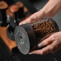 Zwei Hände halten die Dose seitlich, der Deckel mit rundem Display und zwei Tasten zur Kamera