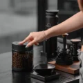 Hand greift nach dem Deckel der Vakuumdose auf einer dunklen Kaffeebar, dahinter eine Mühle und eine Tamperstation
