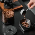 Kaffeebohnen fallen aus einem Löffel in eine Dosierschale auf einer Waage, daneben die offene Dose und ihr Deckel