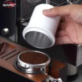 Hand hebt den weißen Dosierbecher von einem Siebträger mit Kaffeemehl in einer Holzstation, innen Metall sichtbar