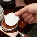 Hand legt een wit papieren schijfje op een portafilter met getampte koffie in een houten station
