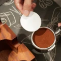 Bovenaanzicht van een hand met een papieren schijfje boven een portafilter met getampte koffie op een bedrukte mat
