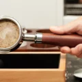 Hand houdt een portafilter met het gebruikte papier op de puck boven de bak van een licht houten knock box