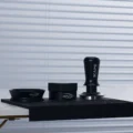 Schwarzer Trichter, Distributor und Tamper auf der schwarzen Matte an der Kante einer Arbeitsplatte, dahinter ein Rollo