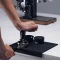 Hand tampt mit dem schwarzen V3 Tamper in einen Siebträger auf der schwarzen Matte, dahinter Trichter und Distributor