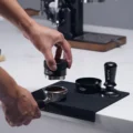 Hand senkt den schwarzen Distributor auf einen Siebträger mit Kaffeemehl auf schwarzer Matte, dahinter Trichter und Tamper
