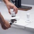 Hand senkt den weißen Distributor auf einen Siebträger mit Kaffeemehl auf der weißen Matte, daneben Trichter und Tamper