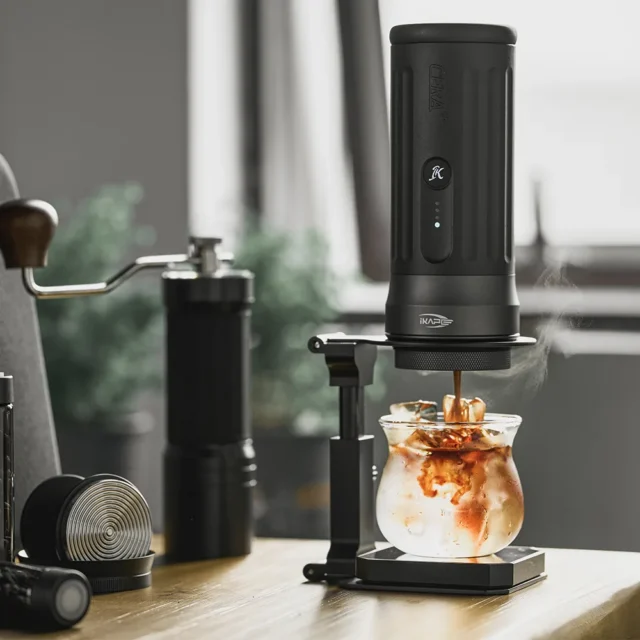 Ständer hält eine tragbare Espressomaschine beim Brühen über einem Glas mit Eis, daneben eine Handmühle