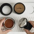 Draufsicht: Eine Hand zeigt die gerillte Stahlfläche des Tampers neben einem Sieb mit in Ringen getamptem Kaffee