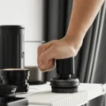 Hand drückt den schwarzen Impact-Tamper mit der ganzen Handfläche in ein Sieb, auf einer weißen gerippten Matte