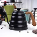 Schwarze Schwanenhalskanne auf weißer Arbeitsfläche, dahinter unscharf weitere Kannen und Kaffeezubehör