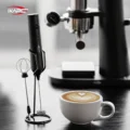 Aufschäumer in seinem schwarzen Drahtständer neben einem Latte mit Herz als Latte Art, dahinter eine Espressomaschine