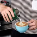 Hand gießt Latte Art aus dem dunkelgrünen Kännchen ohne Griff in eine hellblaue Tasse