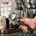 Hand hält den IKAPE Hantom Bottomless-Siebträger vor einer verchromten Espressomaschine, dahinter Kännchen und Tamper