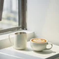 Weißes IKAPE Kännchen neben einer Tasse mit Latte Art auf einer sonnigen Fensterbank