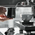 Hand hält das schwarze Kännchen am Hohlgriff neben einer Dampflanze, auf der Abtropfschale eine Tasse auf einer Waage