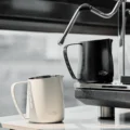 Schwarzes und weißes IKAPE Kännchen, das schwarze unter der Dampflanze einer Espressomaschine, das weiße davor