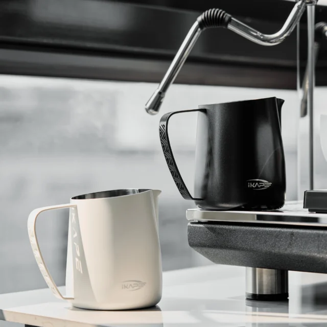 Schwarzes und weißes IKAPE Kännchen, das schwarze unter der Dampflanze einer Espressomaschine, das weiße davor