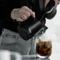 Milch aus dem schwarzen Kännchen fließt in ein Glas Eiskaffee, in der anderen Hand ein Milchaufschäumer