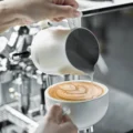 Weißes Kännchen gießt ein Blattmuster in eine weiße Tasse vor einer Espressomaschine