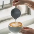 Nahaufnahme: Das weiße Kännchen gießt Latte Art in eine Tasse am Fenster, IKAPE auf dem Griff