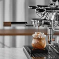 Siebträger mit silbernem Griff in einer Espressomaschine, Espresso läuft in ein Glas mit Eis auf einer Waage