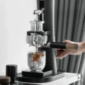 Hand hält den Siebträger mit schwarzem Griff in einer manuellen Espressomaschine, Espresso tropft in ein Glas mit Eis