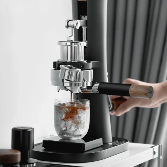 Hand hält den Siebträger mit schwarzem Griff in einer manuellen Espressomaschine, Espresso tropft in ein Glas mit Eis